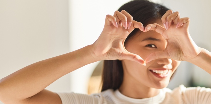 A woman creating a heart with her hands around her eye.]