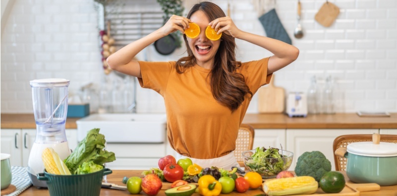A woman holding orange fruit slices in front of her eyes, with a variety of fruit and vegetables in front of her on the counter