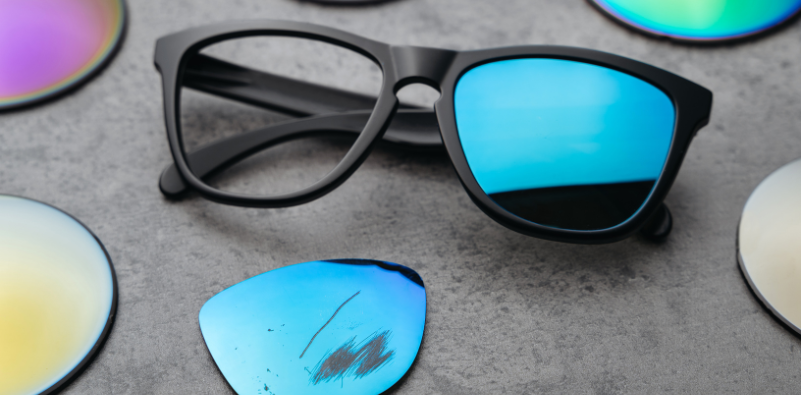 Closeup of broken eyeglasses with damaged colored lens surrounded by different lenses on table.