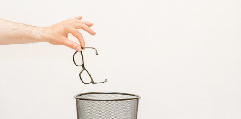 A hand holding a pair of eyeglasses over a trash can.