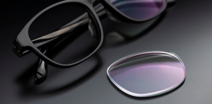 A pair of black eyeglasses with one lens missing, next to a replacement prescription lens.