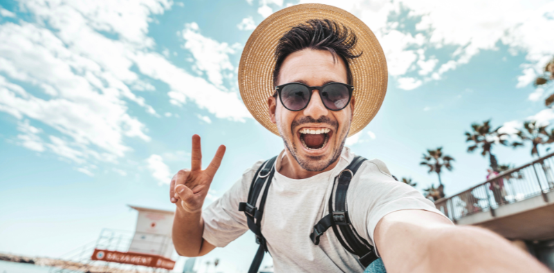 A man wearing a hat and sunglasses taking a selfie at the beach.