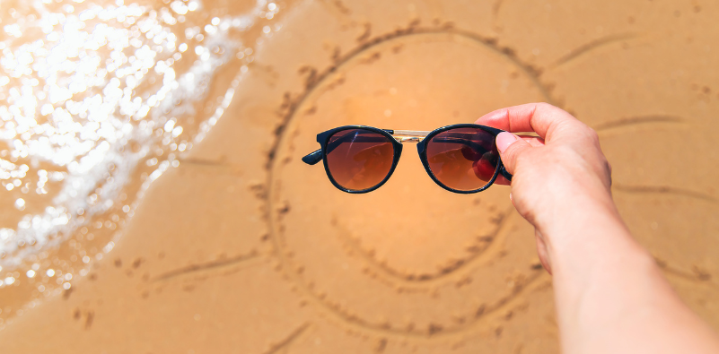 A pair of sunglasses held above a sun drawn in the sand.
