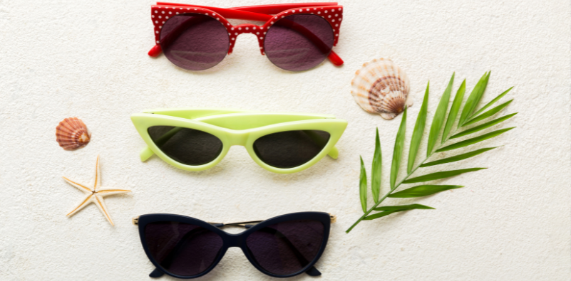 Three different styles of cat eye sunglasses on the sand, surrounded by shells.