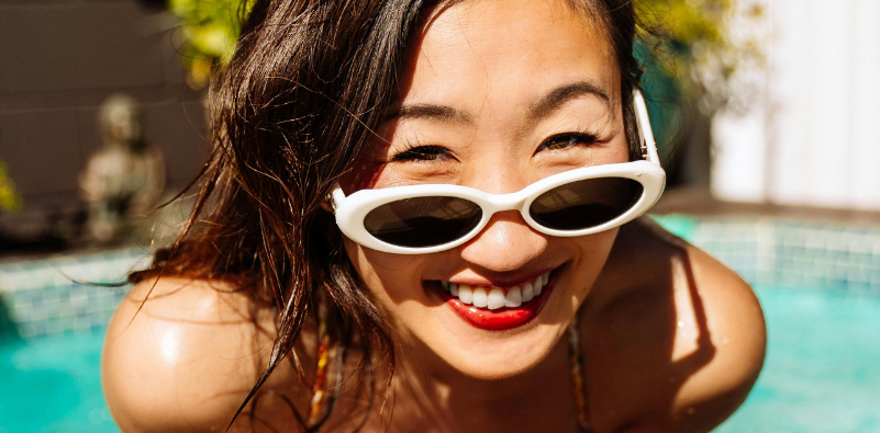 An Asian woman by a swimming pool, wearing a pair of white oval sunglasses on the end of her nose.