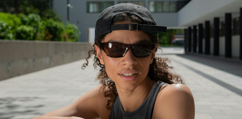 A person wearing sporty, wraparound sunglasses and a backwards cap, outside.