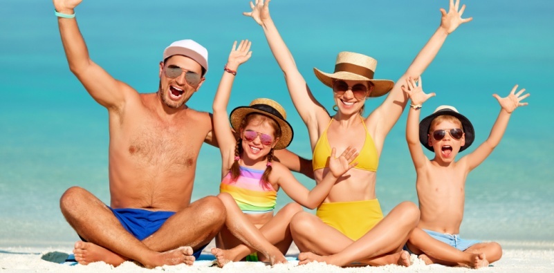 A happy family sat on the beach with their arms up, wearing sunglasses and hats.