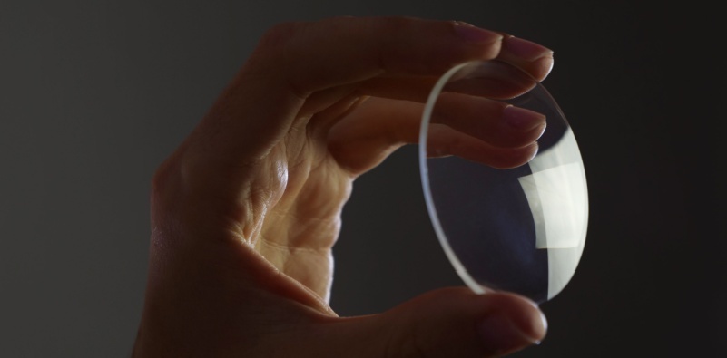 A hand holding up an eyeglasses lens, showing white reflections.