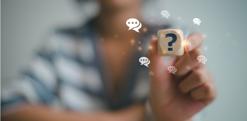 – A blurred out person holding a wooden dice with a question mark on it, surrounded by illustrated speech marks
