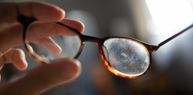 A person holding a pair of eyeglasses with scratched and smudged lenses.