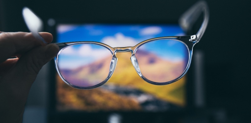 A person holding a pair of blue light blocking glasses in front of a digital screen, emphasizing the importance of eyewear for screen-heavy lifestyles