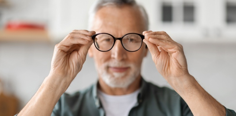 A senior man holding his glasses away from his face, as if he is about to put them on.