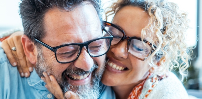 An adult couple in their 40’s wearing glasses, smiling and embracing each other.