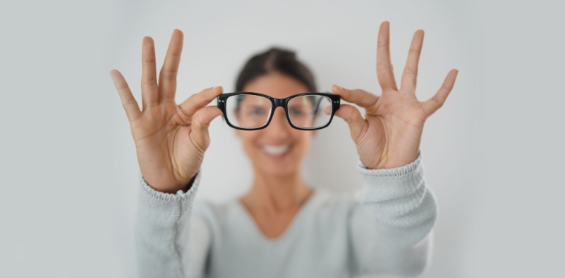 A brunette woman, with her arms outstretched holding a pair of black eyeglasses.