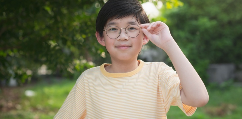 A child with myopia wearing a pair of round eyeglasses, stood outside in a garden.