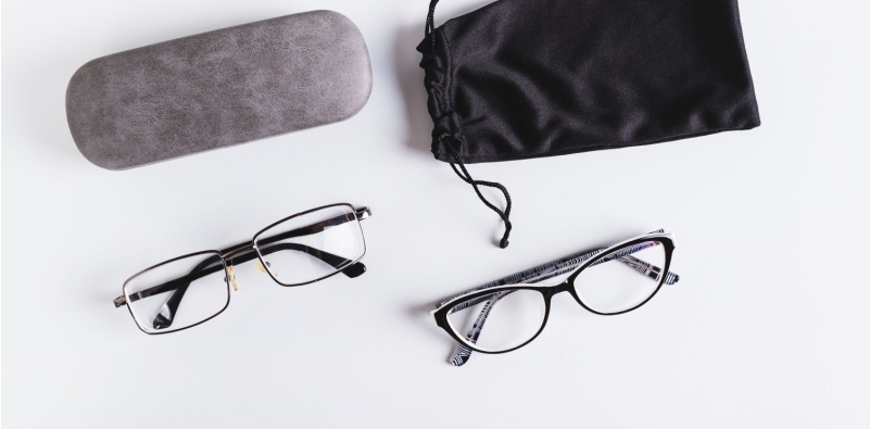 A top view of two pairs of eyeglasses on a white background alongside a hard and soft eyeglasses case.
