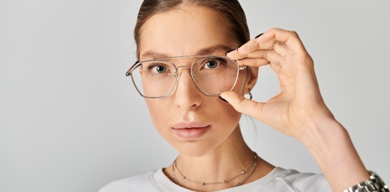A young woman confidently wearing pilot style glasses and a white shirt against a grey background.