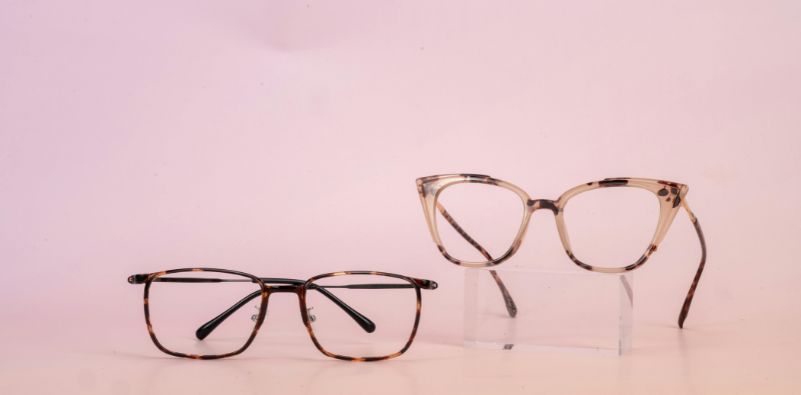 Two pairs of tortoiseshell/mottled colored eyeglasses on a pink background