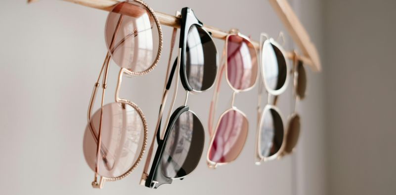 A range of different style eyeglasses with colorful tinted lenses, hanging on a wooden coat hanger.